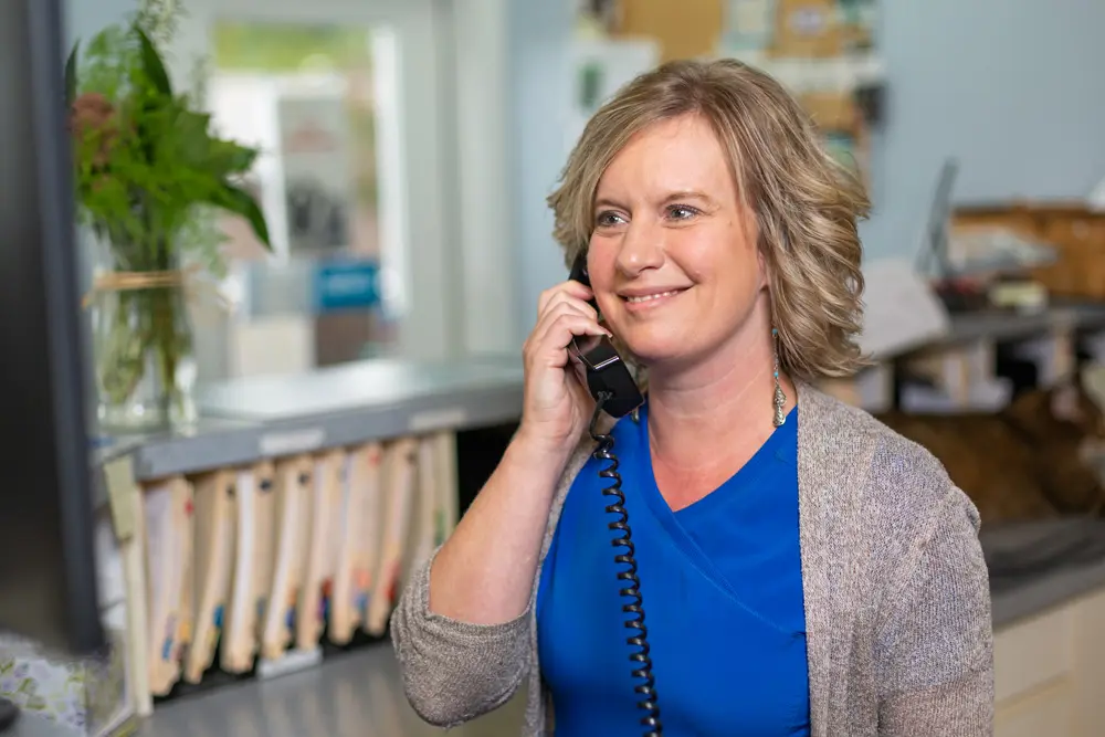 Smiling receptionist answering phone at veterinary clinic front desk with patient files and plant in background.