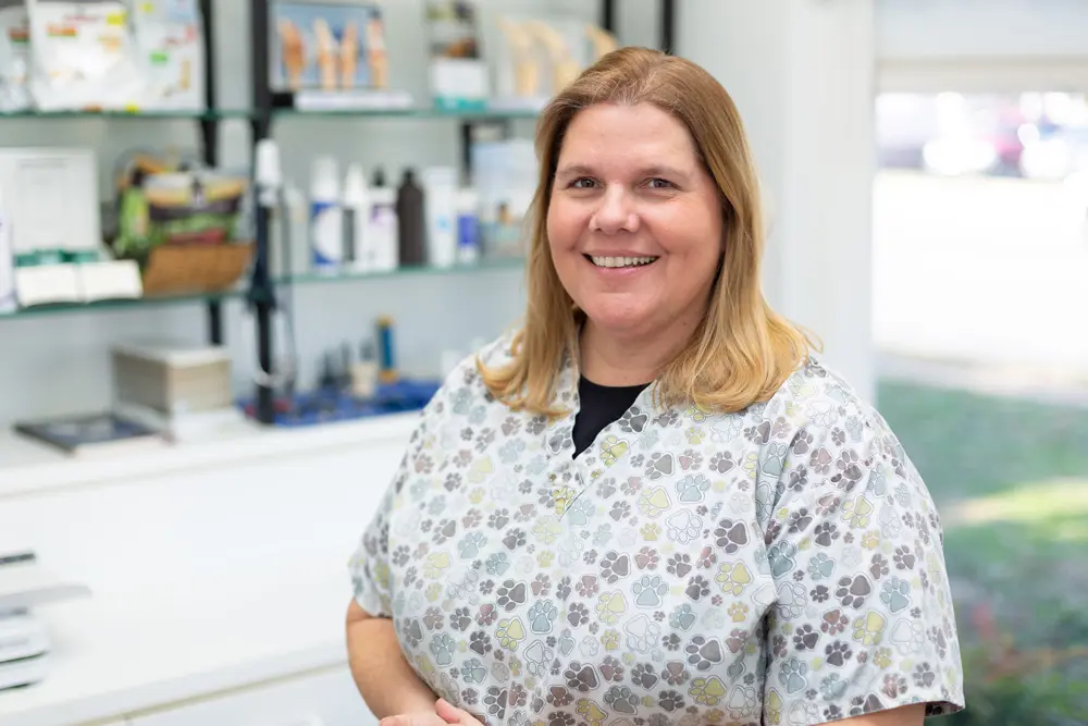 Smiling veterinary clinic staff member in paw-print scrubs standing in a bright exam room with medical supplies on shelves behind.